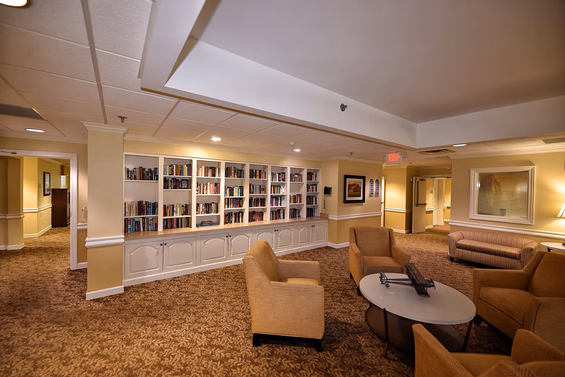 Cozy senior living lounge with beige seating, round table, and wall of built-in bookshelves.