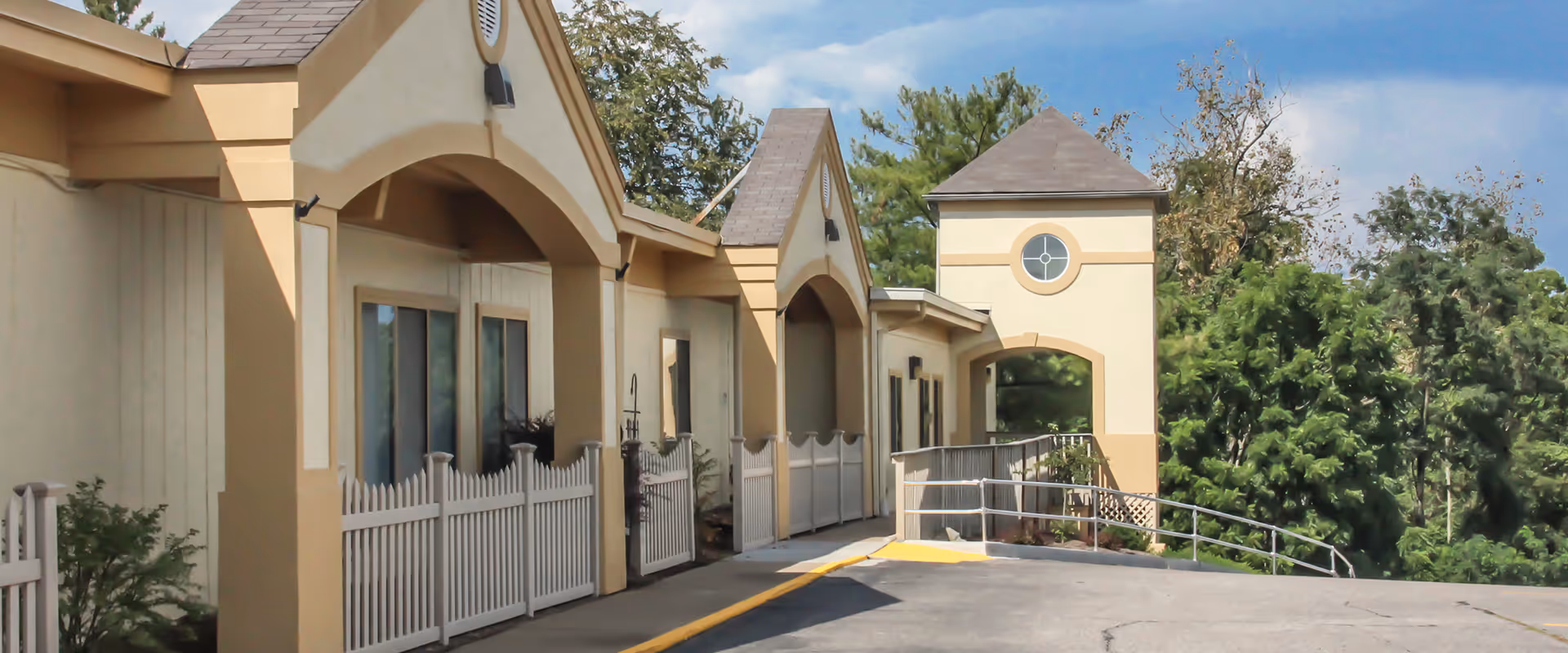 Exterior view of a beige and tan building with arched doorways and a small tower-like structure. There is a white picket fence along the walkway and greenery in the background under a partly cloudy sky.