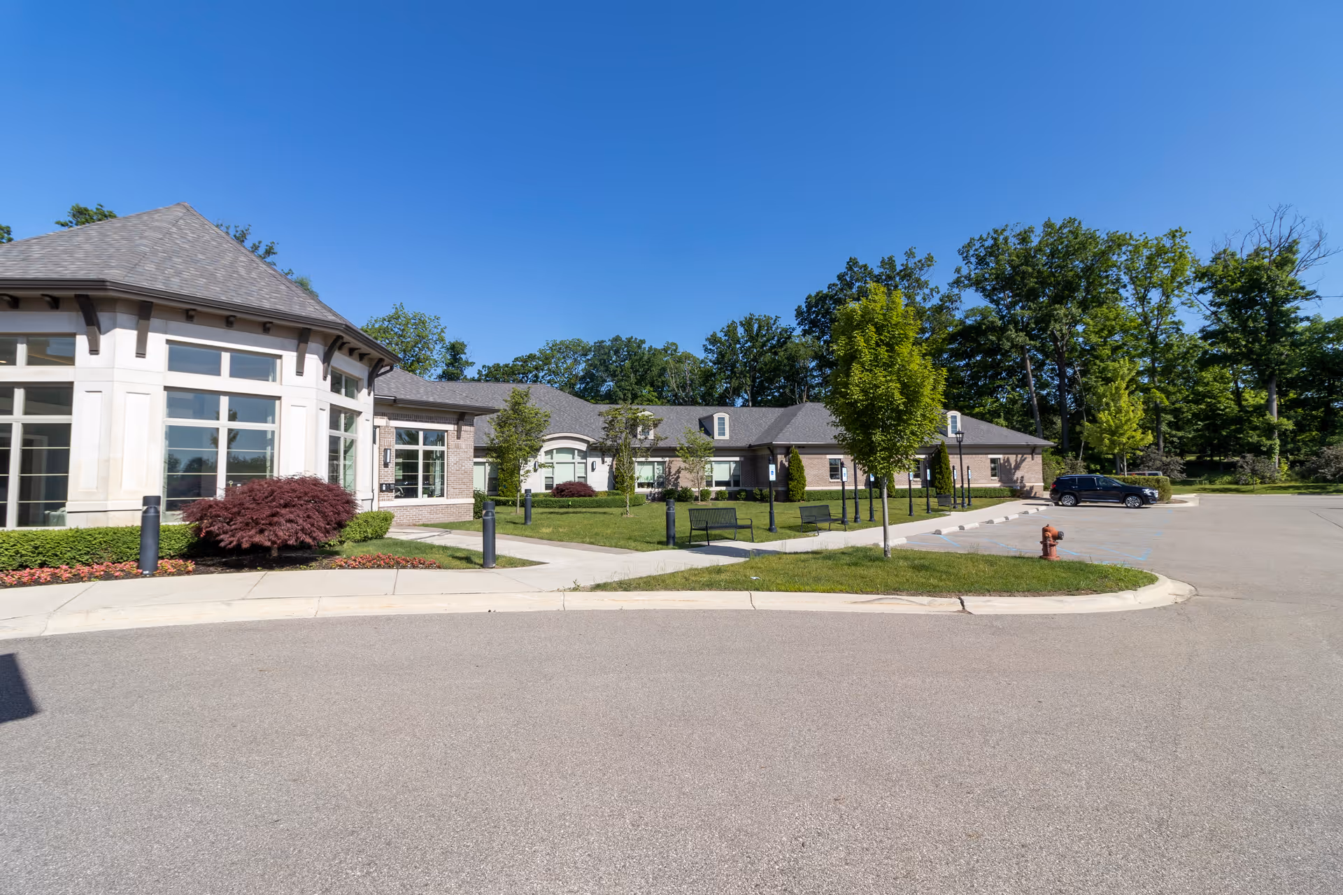 Front exterior of a single-story senior living facility with large windows, landscaped lawn, benches, and a parking area under a clear blue sky.
