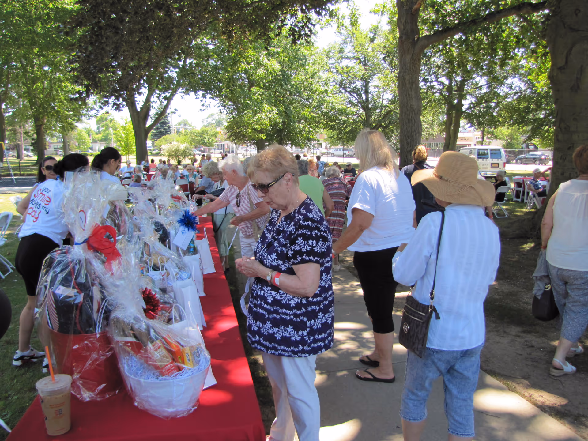 Outdoor park setting with seniors and staff examining gift baskets on a red table.