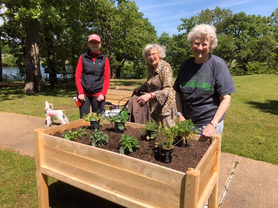 Three elderly women outdoors standing around a raised wooden garden bed with small plants in pots ready for planting. One woman is wearing a red cap and black vest, another is in a leopard print jacket, and the third is in a dark t-shirt. There is a small white dog on a leash nearby and green trees and grass in the background under a clear blue sky.