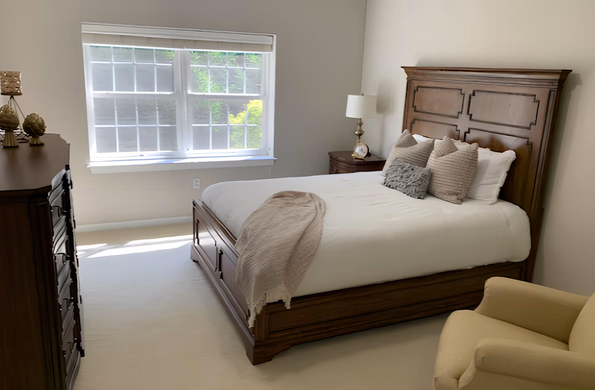 A bedroom with a wooden bed frame and headboard, neatly made with white bedding and several decorative pillows. A beige throw blanket is draped over the foot of the bed. There is a wooden dresser on the left side with decorative items on top, a nightstand with a lamp and clock on the right side, and a beige armchair in the foreground. A large window with white blinds lets in natural light.