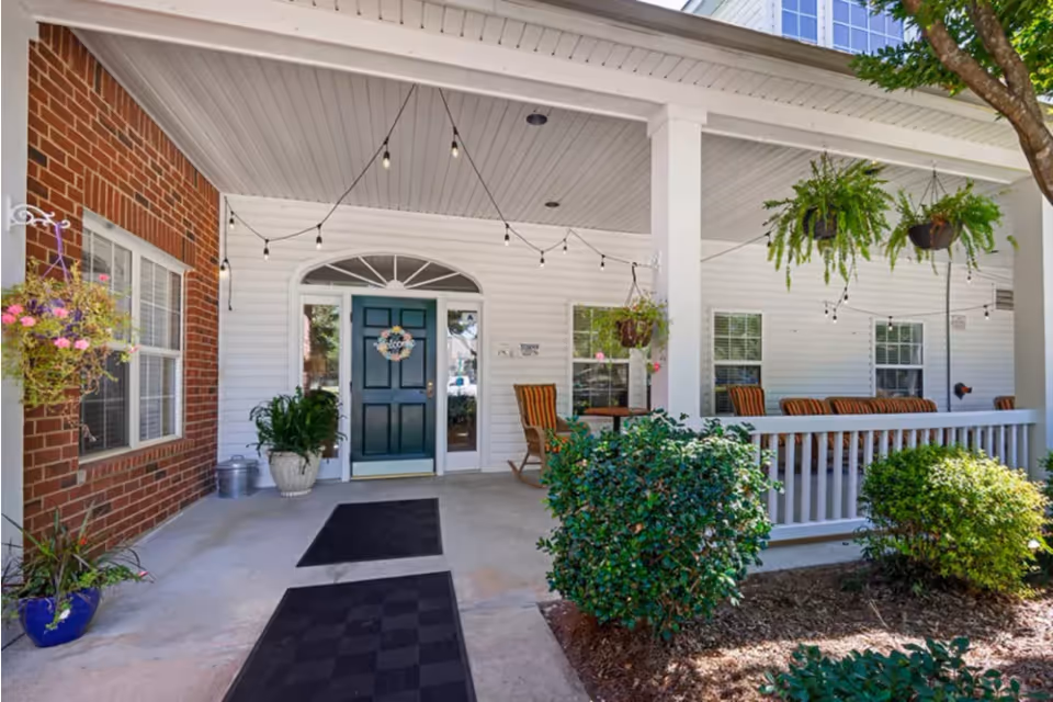 Front porch entrance of a senior living facility with a dark green door decorated with a welcome wreath, white siding, brick wall on the left, hanging plants, string lights, outdoor seating with striped cushions, and neatly trimmed bushes in the foreground.