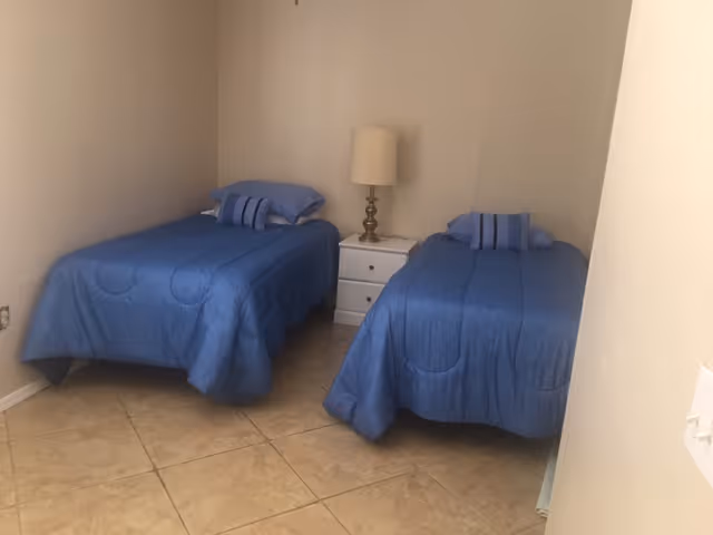 A small bedroom with two twin beds covered in blue bedspreads and matching pillows. Between the beds is a white nightstand with a lamp on top. The room has beige tiled flooring and plain light-colored walls.