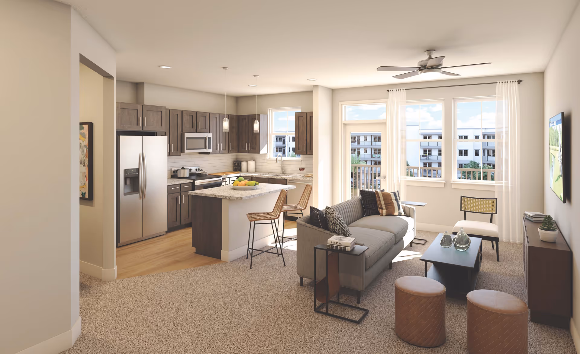 A bright and modern open-concept living room and kitchen area in a senior living facility. The living room features a gray sofa with multiple pillows, a small side table with books, two round ottomans, a coffee table with decorative glass bottles, a chair, and a wall-mounted TV. The kitchen has dark wood cabinets, stainless steel appliances including a refrigerator and microwave, a kitchen island with a bowl of fruit, and two bar stools. Large windows and a glass door let in natural light and offer a view of nearby buildings.