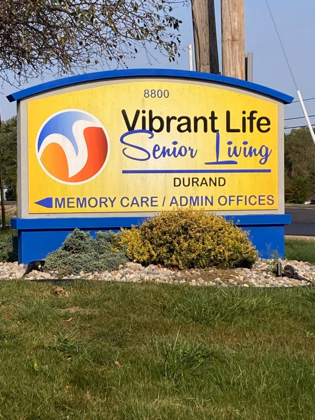 Outdoor sign for Vibrant Life Senior Living in Durand, with directions to Memory Care and Admin Offices, surrounded by grass, bushes, and small rocks.