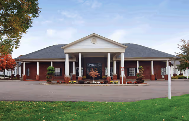 Front view of a single-story red brick senior living building with white columns, a covered entrance, and landscaped grounds.