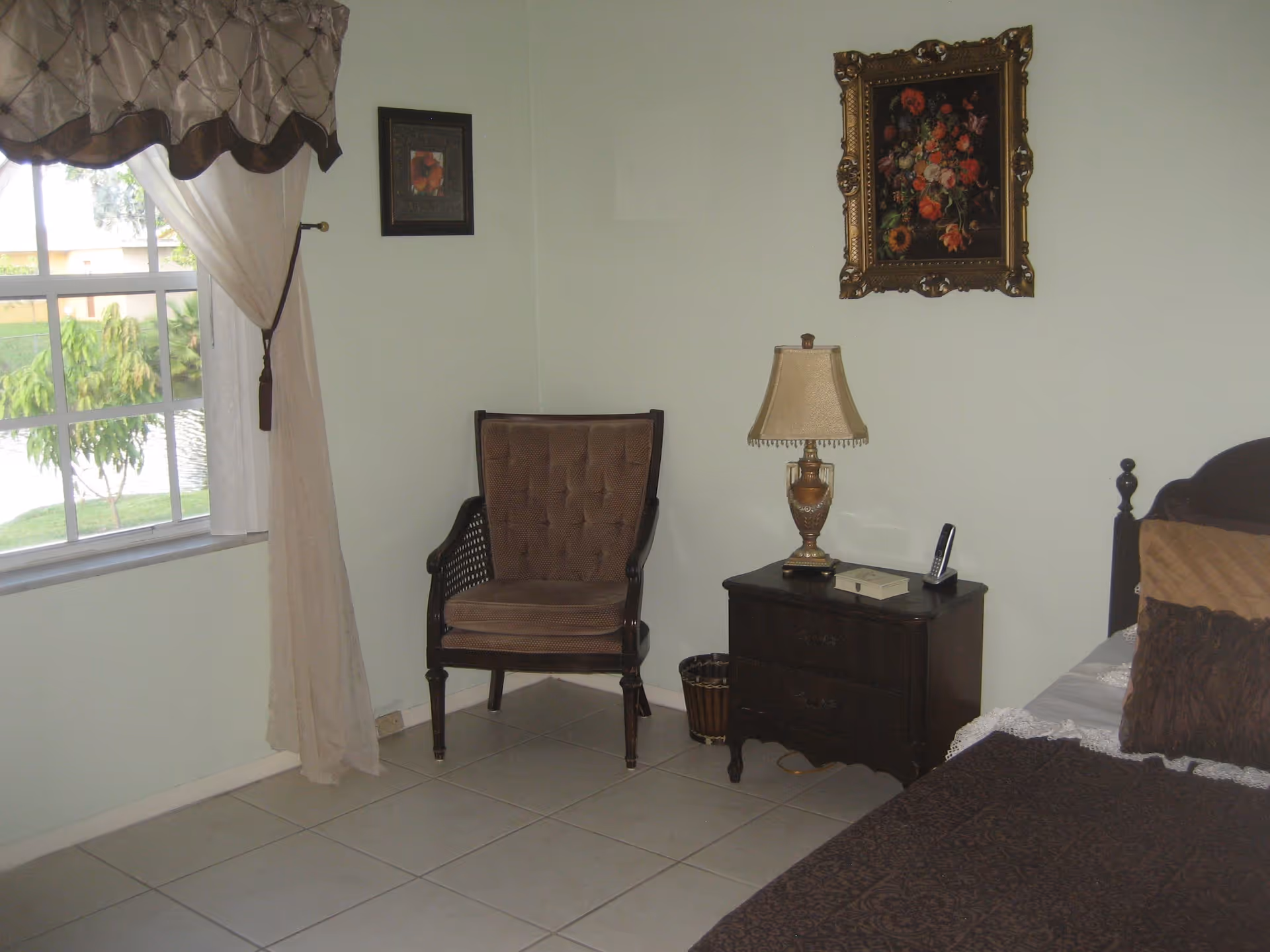 Bedroom corner with an upholstered chair, nightstand and lamp next to a bed beneath framed artwork and a window.