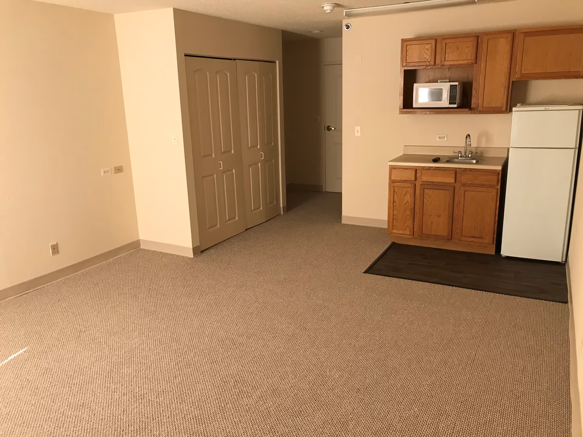 Empty room with beige carpet and walls, featuring a small kitchenette with wooden cabinets, a microwave, a sink, and a white refrigerator. There is a closet with double doors and a closed door in the background.