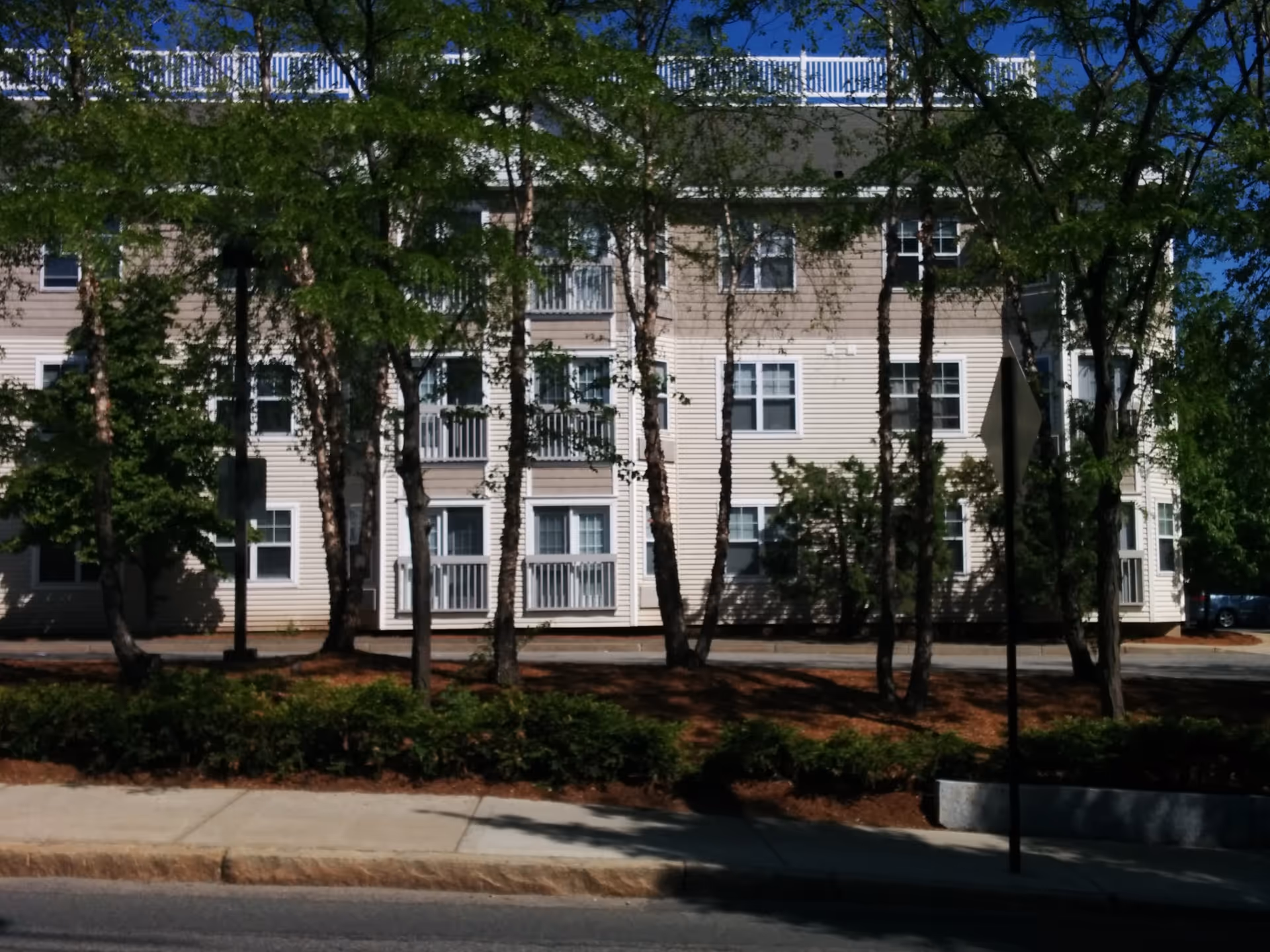 A multi-story beige apartment building with balconies behind trees along a sidewalk.