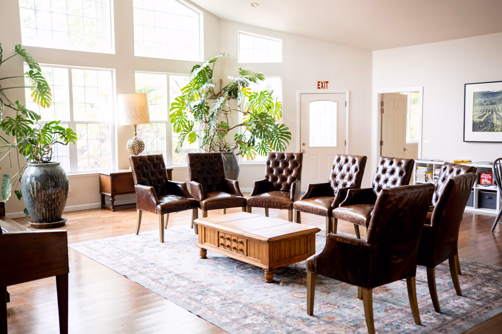 Bright common area with brown leather chairs around a wooden coffee table, large windows and plants.