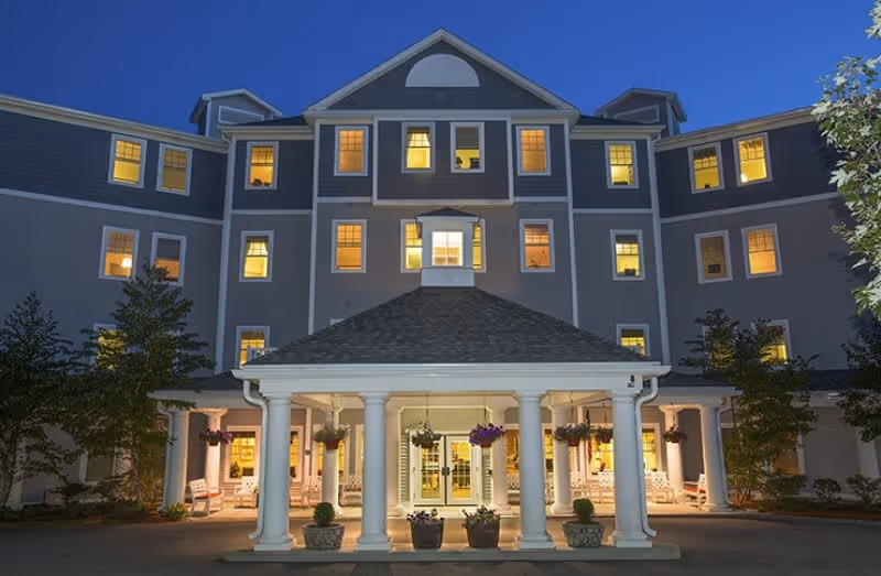 Front entrance of a multi-story senior living building at dusk with a columned portico and lit windows.