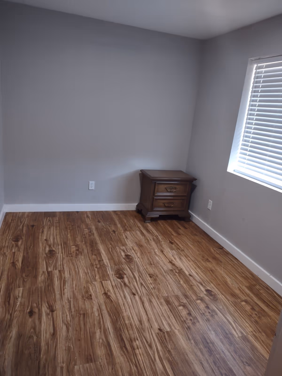 Small empty bedroom with wood-look flooring, gray walls, a window with blinds, and a wooden nightstand in the corner.