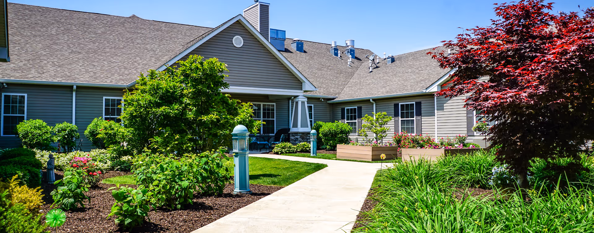 A bright outdoor garden area at Bickford of Crystal Lake featuring a paved walkway surrounded by lush green plants, bushes, and colorful flowers. The building exterior is visible with beige siding, multiple windows, and a sloped roof under a clear blue sky.