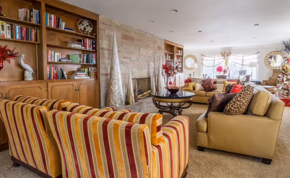 Cozy senior living lounge with striped couches, beige seating, fireplace, bookshelves, holiday decor.