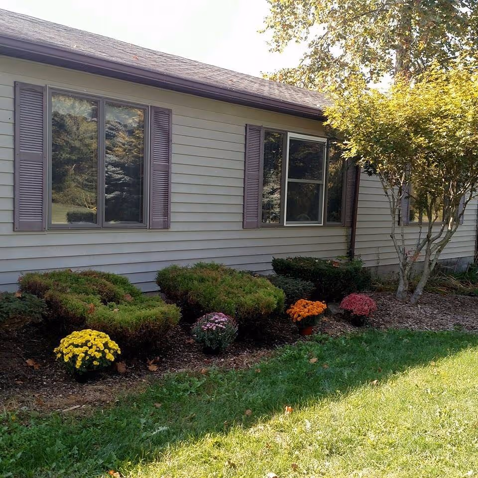 Exterior side view of a building with beige siding and purple shutters on the windows. There are neatly trimmed bushes and colorful flowers planted along the foundation, with a tree on the right side and green grass in the foreground.