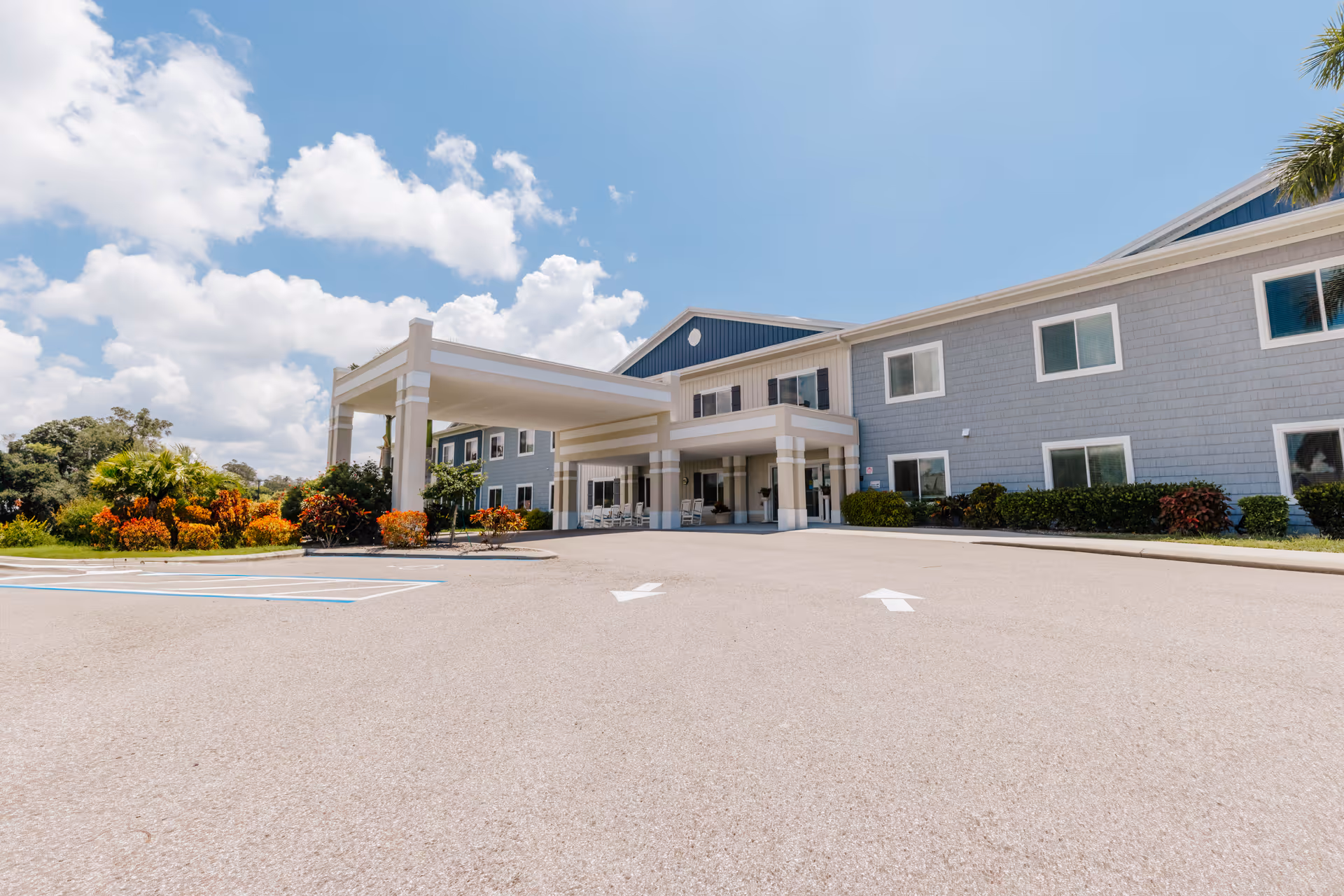 Front entrance of a two-story senior living building with a covered porte-cochère, parking area, and landscaping under a blue sky.