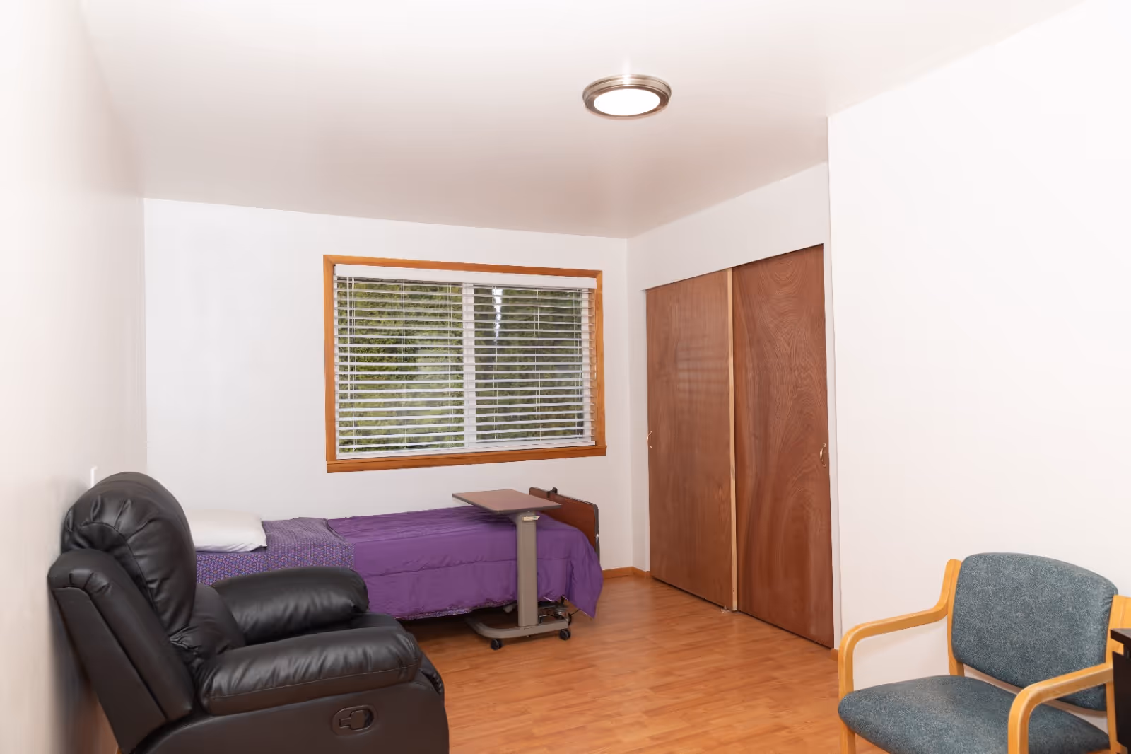 A small bedroom featuring a bed, recliner, window, closet doors, and purple bedding.