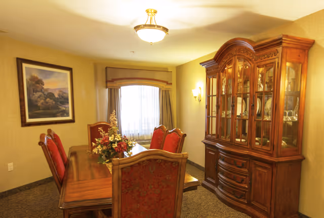 A warmly lit dining room featuring a wooden dining table with six red upholstered chairs. A floral centerpiece decorates the table. On the right side, there is a large wooden china cabinet displaying glassware and dishes. A framed landscape painting hangs on the left wall, and a window with curtains is centered on the back wall.