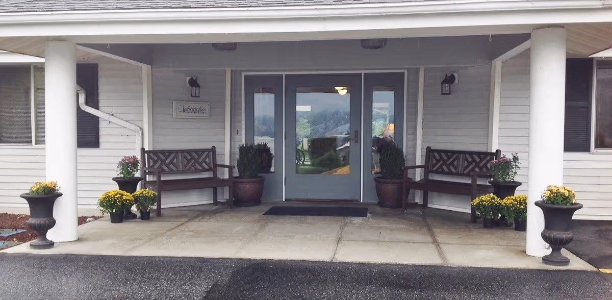 Covered front entrance of a senior living facility with benches and potted plants.