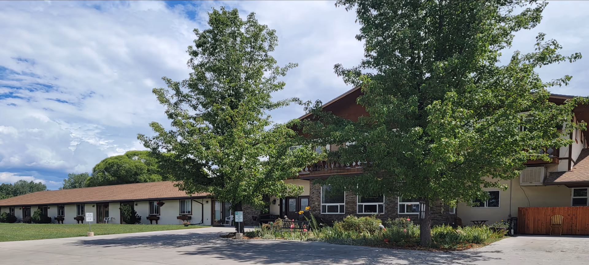Exterior view of Spring Creek Chalet senior living facility showing a two-story building with a brown roof and white walls. There are large green trees and landscaped bushes in front of the building, with a paved driveway and lawn area under a partly cloudy sky.
