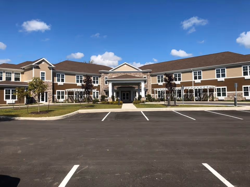 Front exterior of a two-story beige and brown assisted living building with a covered entrance and a parking lot in front.