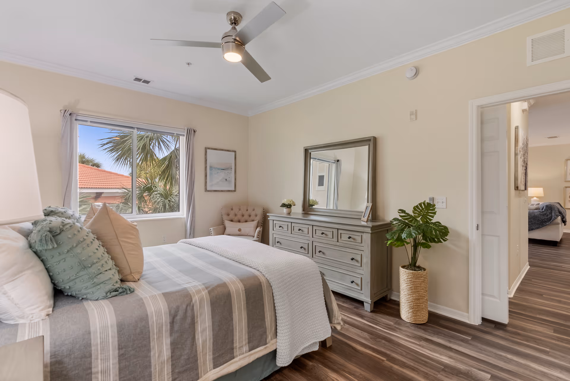 Bright bedroom with a striped bed, dresser and mirror, ceiling fan, and window view.
