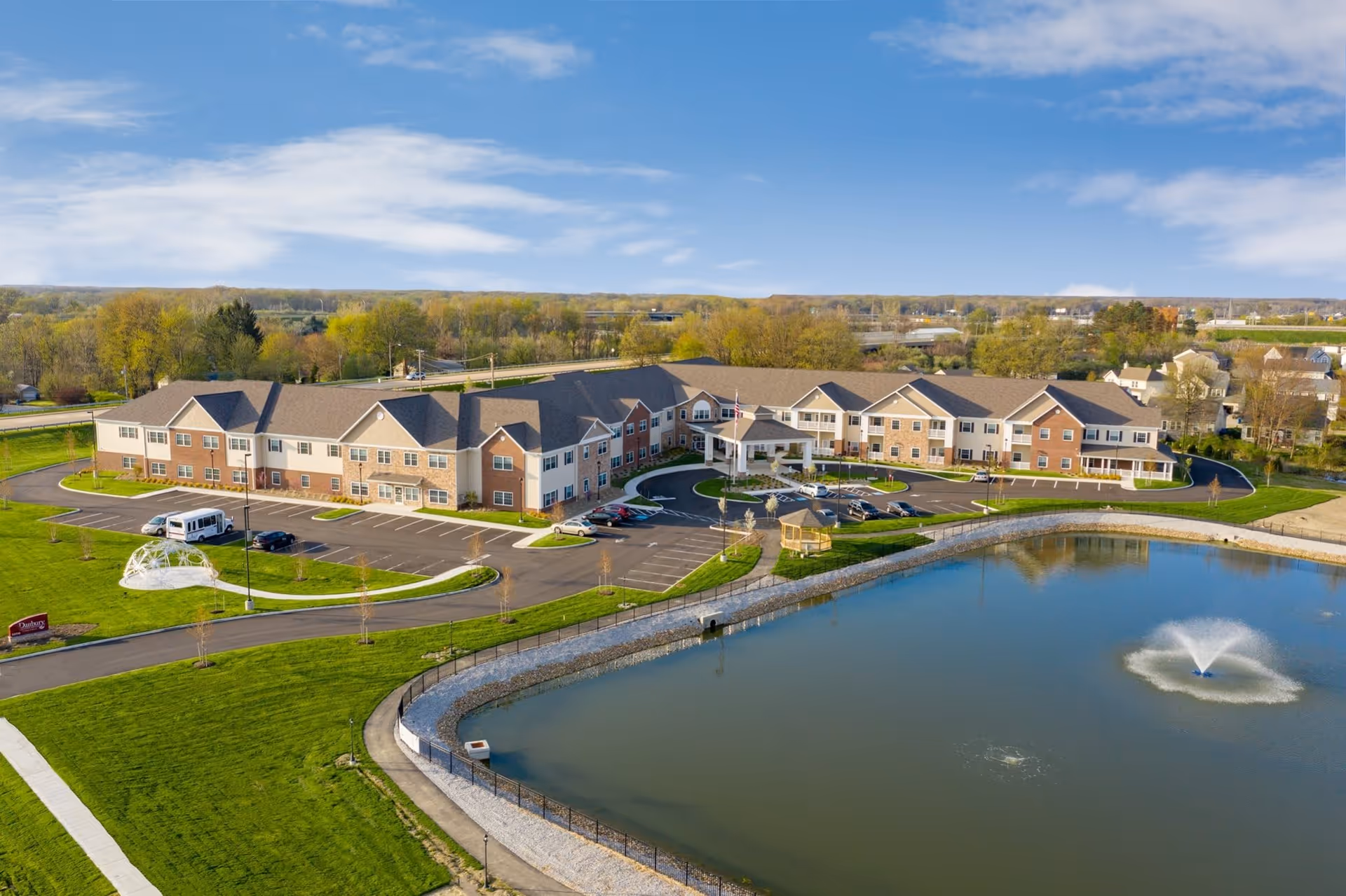 Aerial view of Danbury North Ridgeville senior living facility showing a large two-story building with a parking lot, a pond with a water fountain, green lawns, and a gazebo. The sky is partly cloudy and trees are visible in the background.