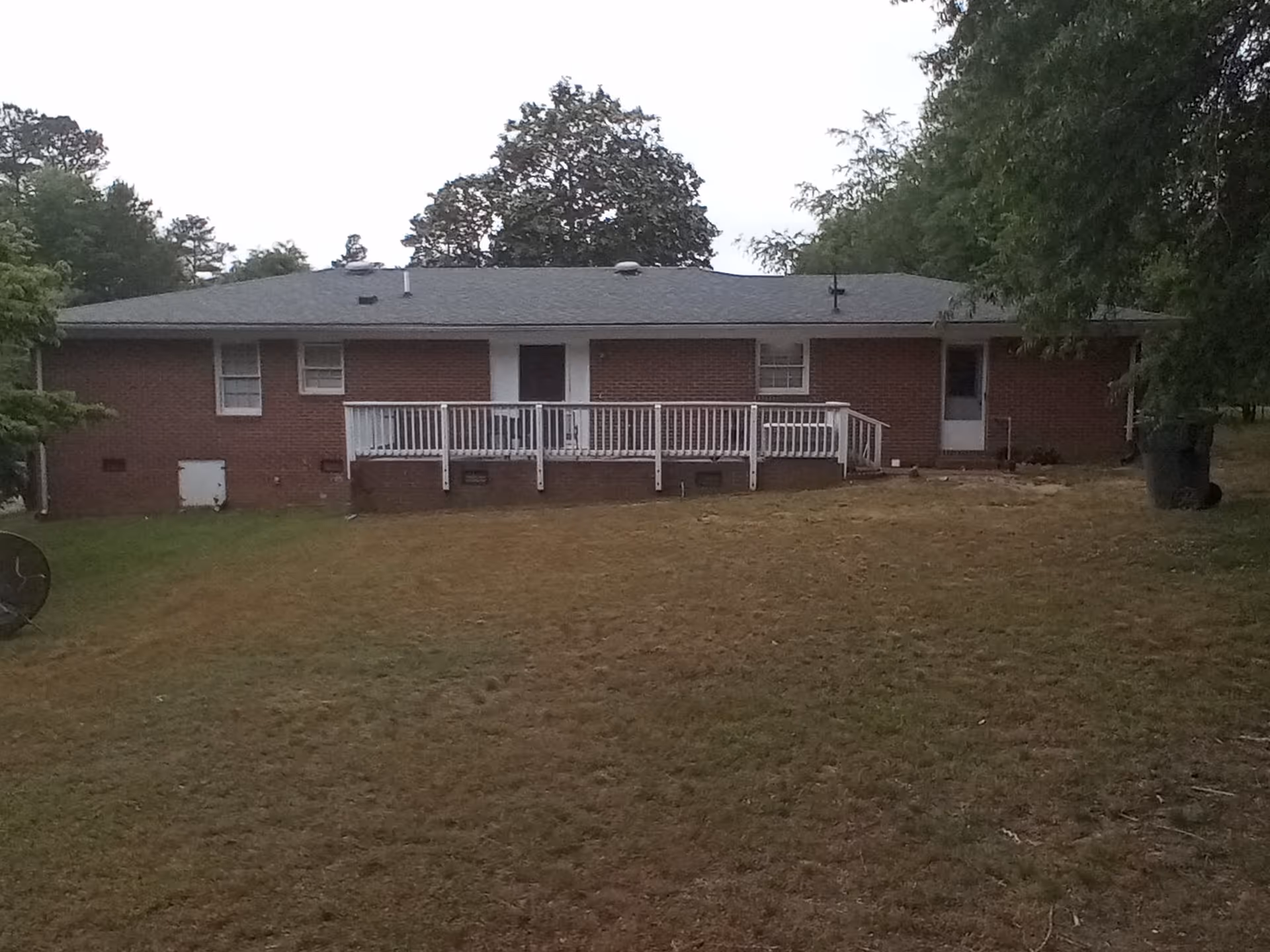 Single-story brick house with a white-railed wooden deck and a large grassy yard under an overcast sky.