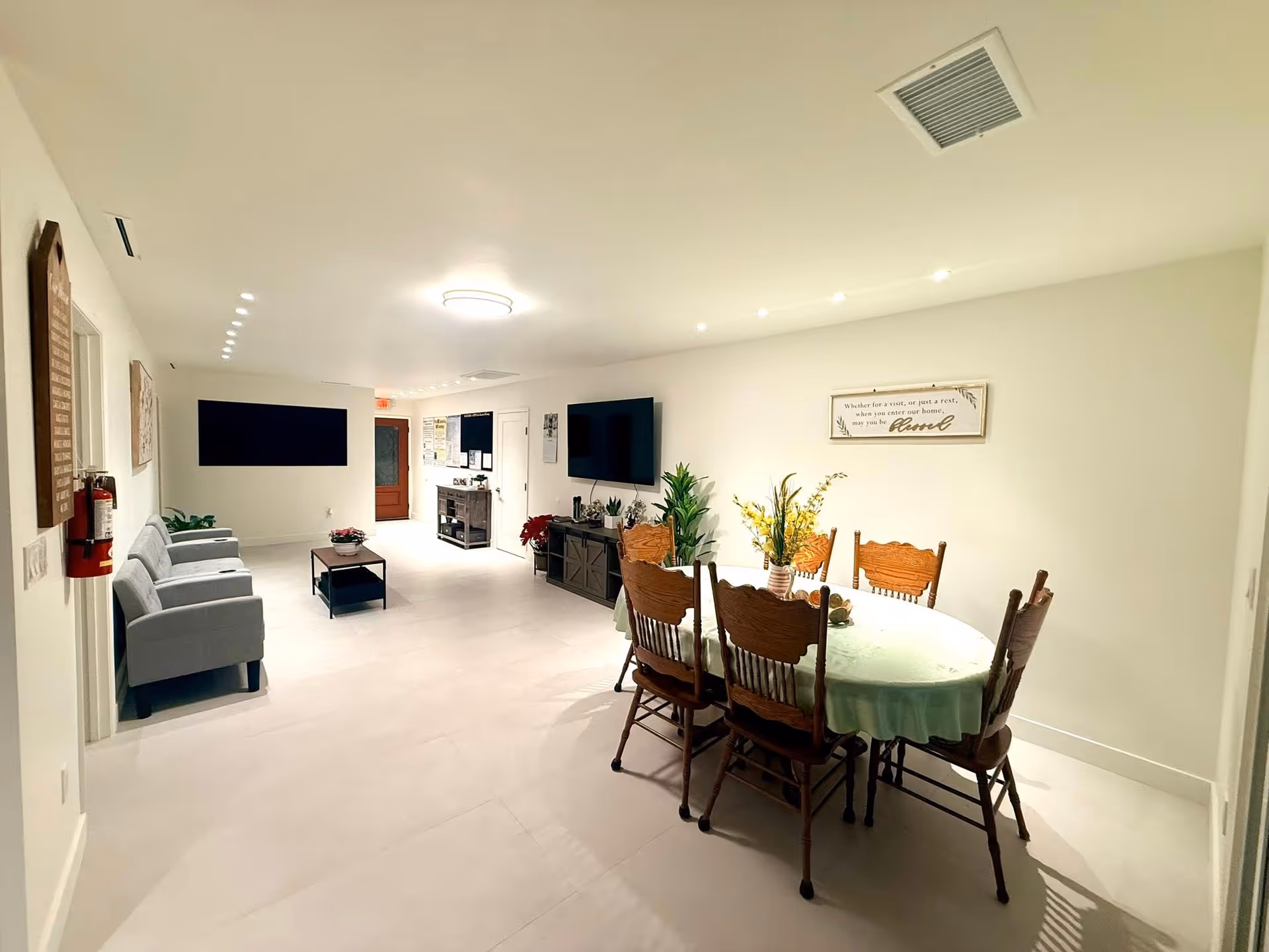 Bright senior living lounge with grey seating, a round dining table, and TVs.