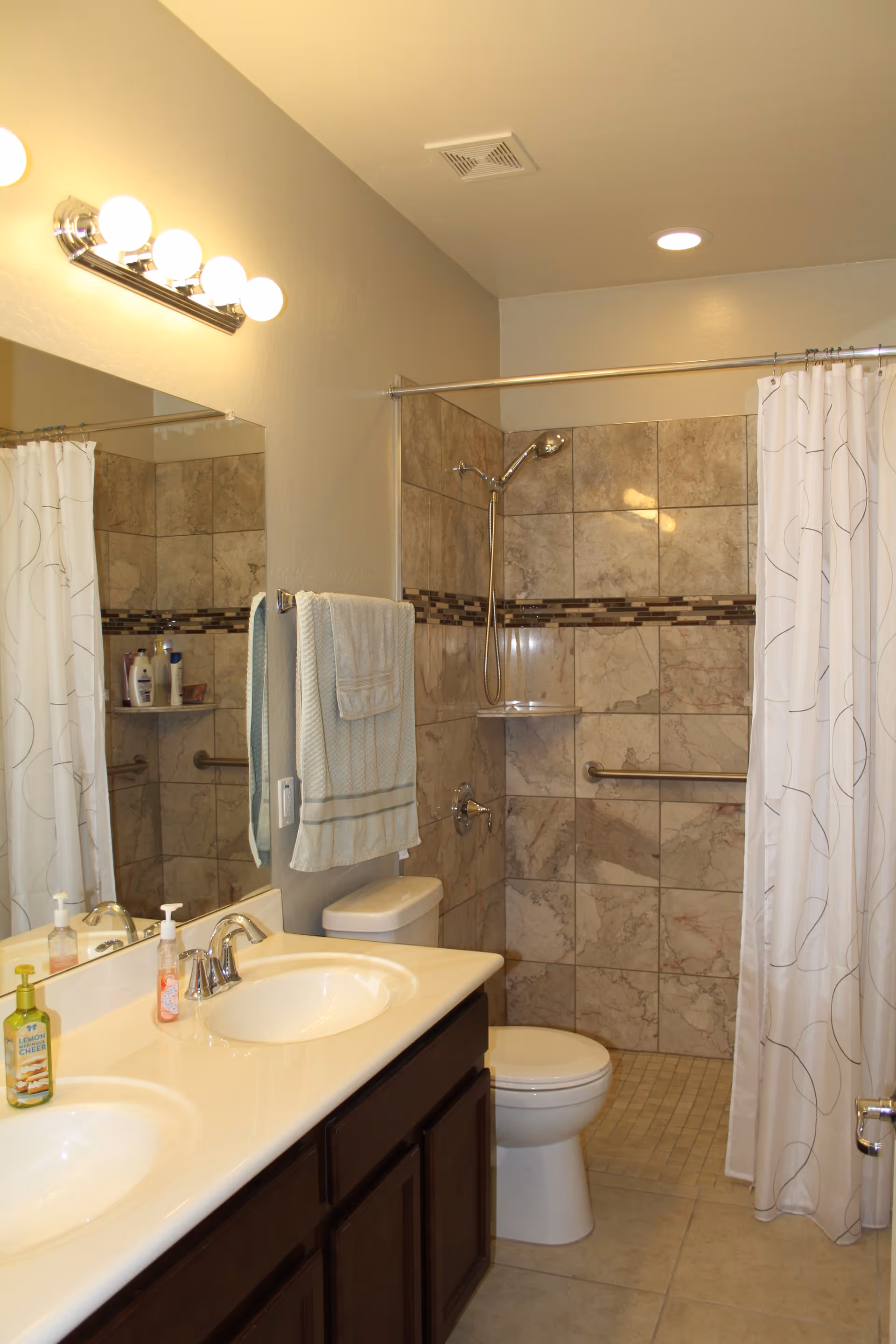 A clean bathroom with a double sink vanity featuring chrome faucets and soap dispensers. Above the sinks is a large mirror with a light fixture containing five round bulbs. Next to the vanity is a toilet, and beyond that is a walk-in shower with beige and brown tiled walls, a chrome showerhead, a grab bar, and a white shower curtain with a subtle pattern. A light blue towel hangs on a towel rack beside the toilet.