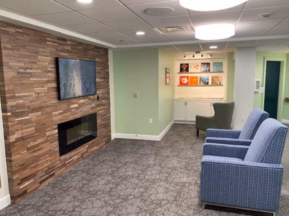 A cozy indoor lounge area with two blue upholstered armchairs facing a wall-mounted flat screen TV above a modern electric fireplace set in a wooden accent wall. The room has light green walls, a patterned carpet, and a small nook with white cabinets and shelves displaying colorful vinyl record covers. A single green armchair is placed near the nook, and the ceiling has recessed lighting and a round ceiling light fixture.
