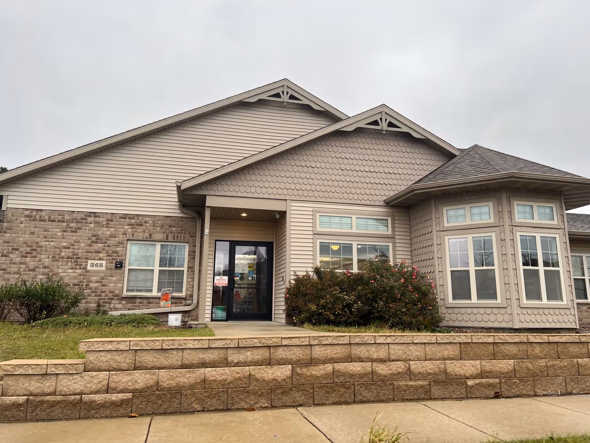 Front entrance of a single-story senior living building with beige siding, brick accents, a glass door, and a low retaining wall.
