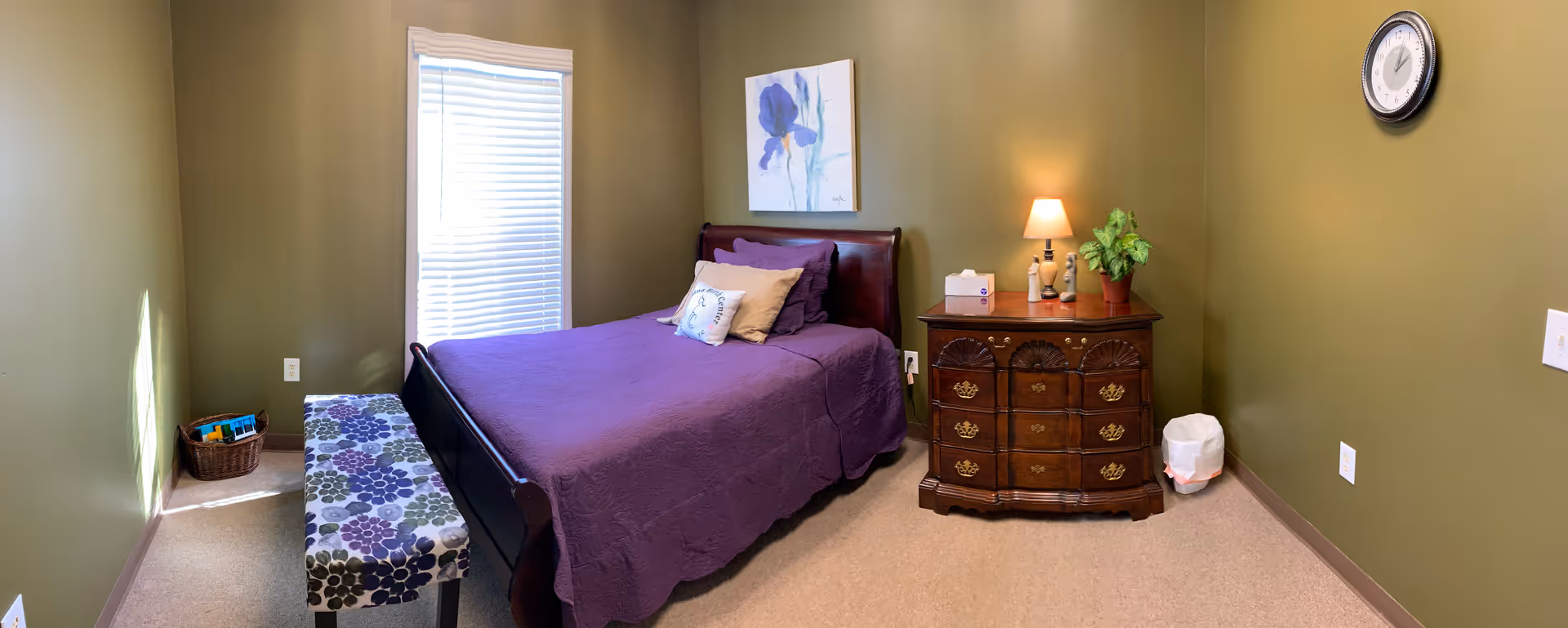 Green walls with a purple bedspread, wooden dresser, bench, plant, and a lamp.