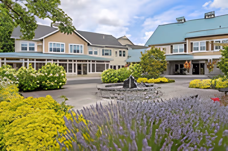 Exterior view of a retirement community building with a blue roof, large windows, and landscaped gardens featuring yellow and purple flowers in the foreground under a partly cloudy sky.