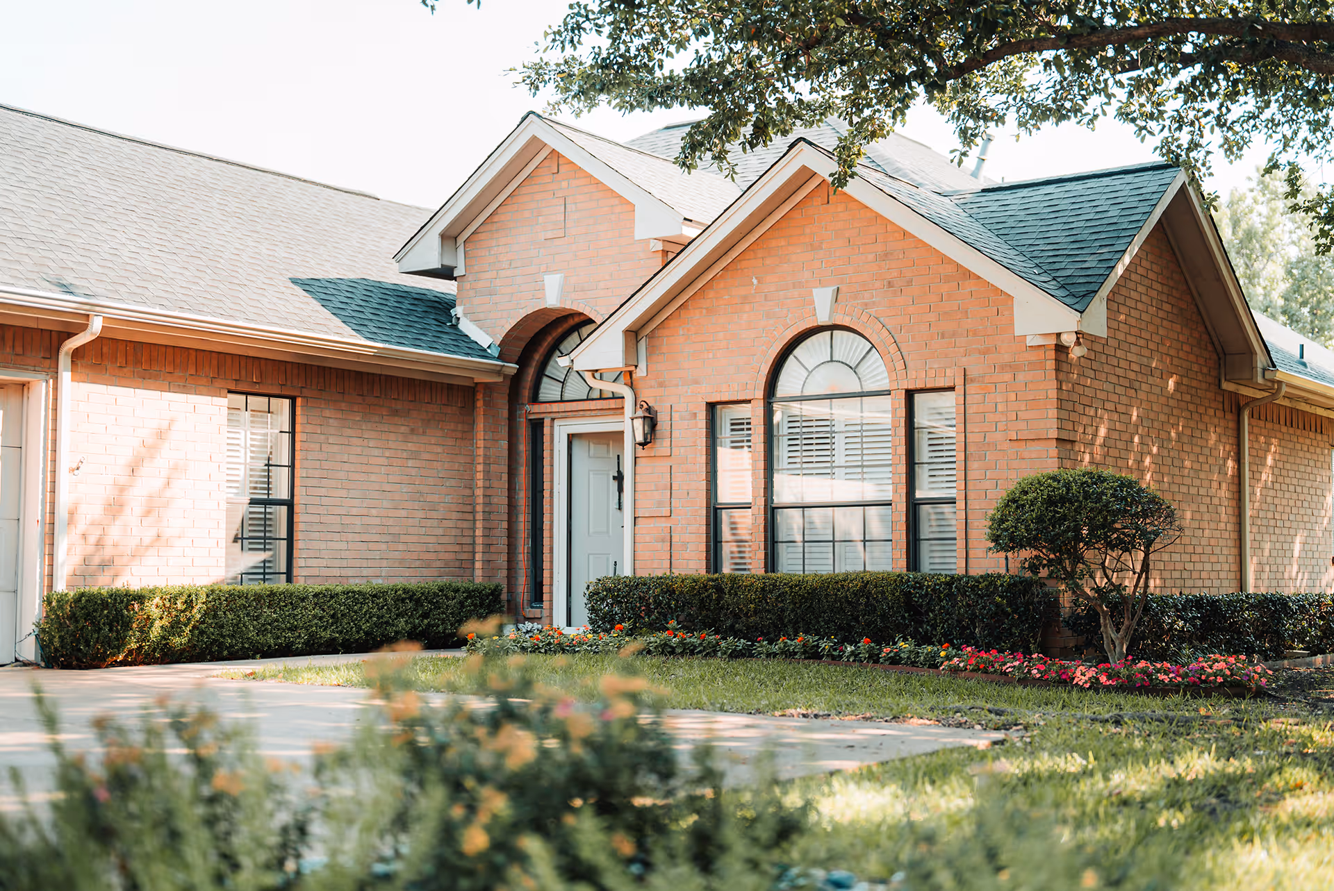 Exterior view of a single-story brick building with a gabled roof, featuring a white front door and large arched windows. The building is surrounded by neatly trimmed bushes, colorful flower beds, and a well-maintained lawn with trees providing shade.