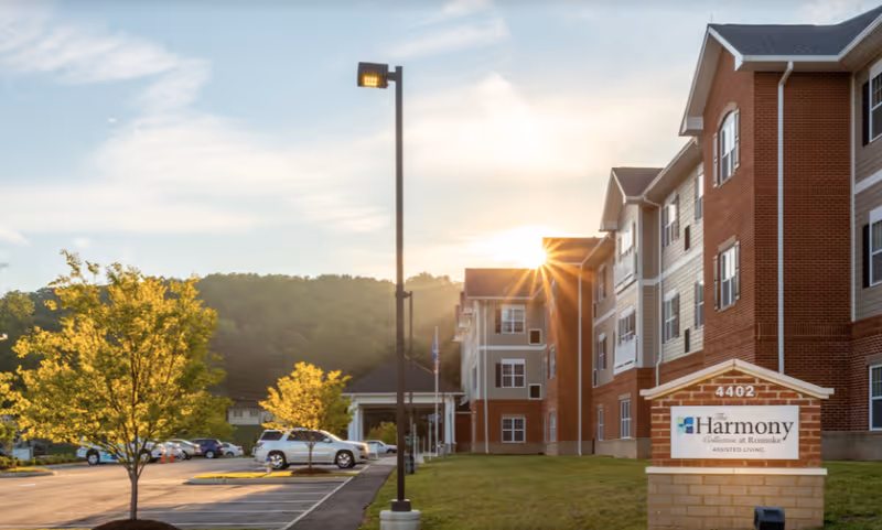 The front exterior of The Harmony Collection at Roanoke assisted living building with a parking lot, trees, and a sign at sunset.