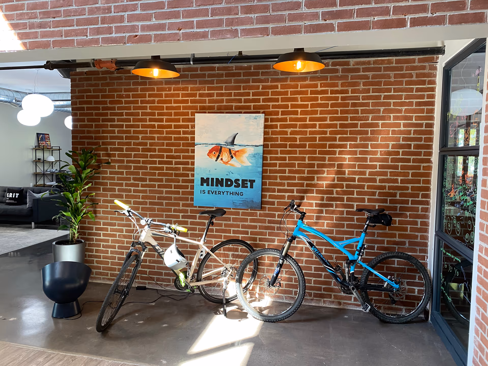 Two bicycles lean against a brick wall beneath a motivational poster in a modern indoor lobby.