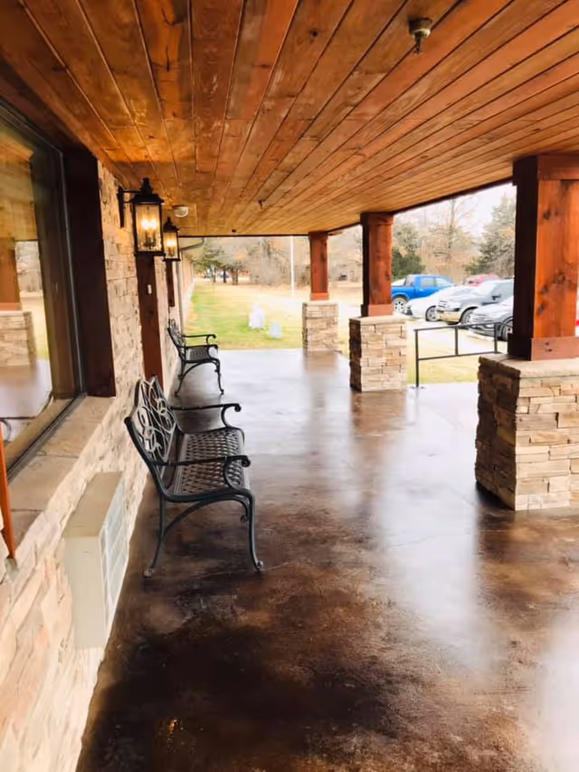 Covered outdoor porch area with a wooden ceiling, stone pillars, and metal benches along the wall. There are wall-mounted lantern-style lights and a view of parked cars and trees in the background.