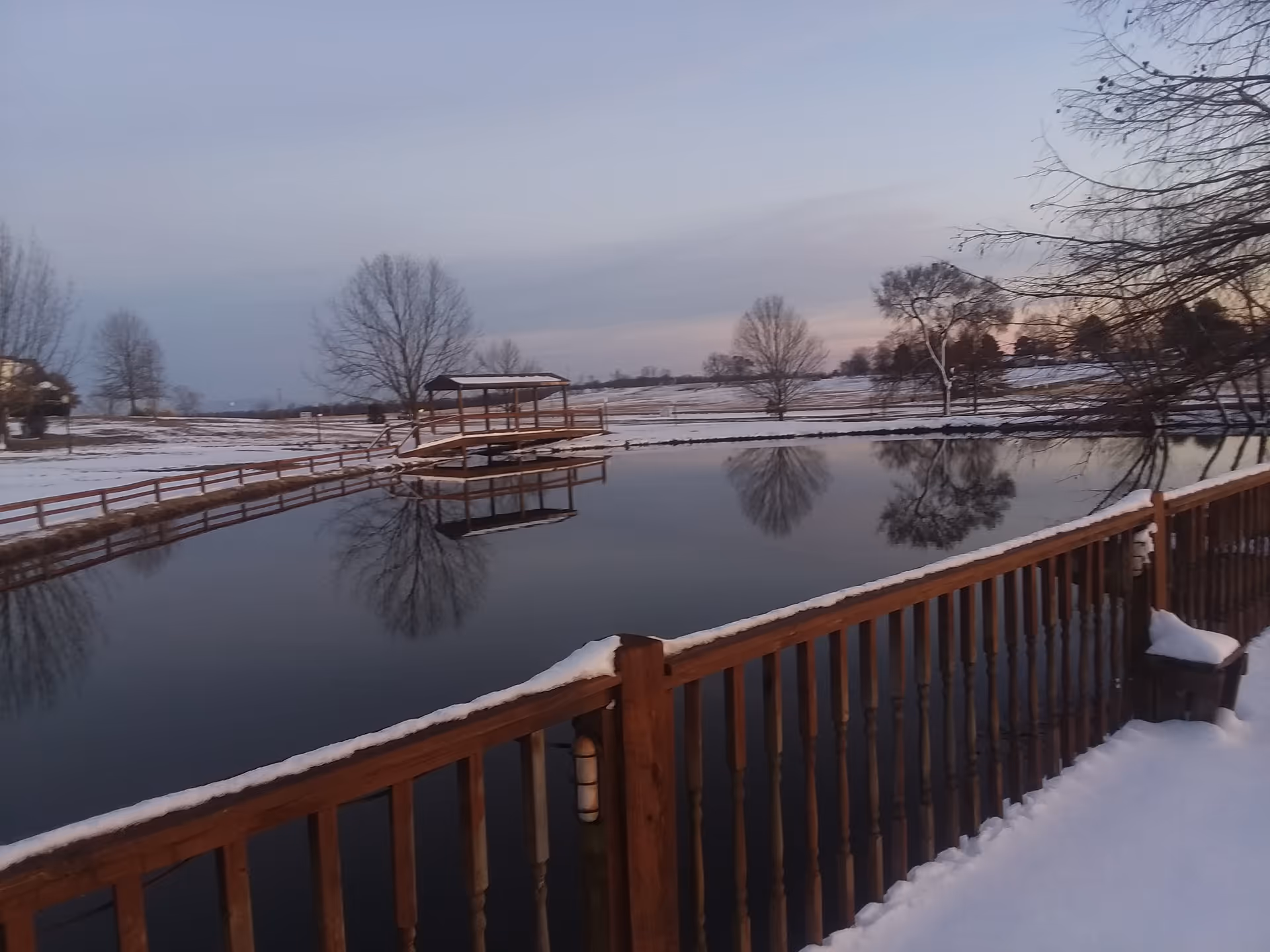 Snow-covered outdoor pond with wooden railing, gazebo, and leafless trees.