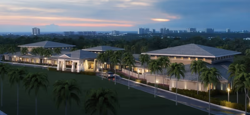 Exterior view of a large, single-story senior living facility building with multiple peaked roofs, surrounded by palm trees and a driveway leading to the entrance, set against a backdrop of a city skyline at sunset.