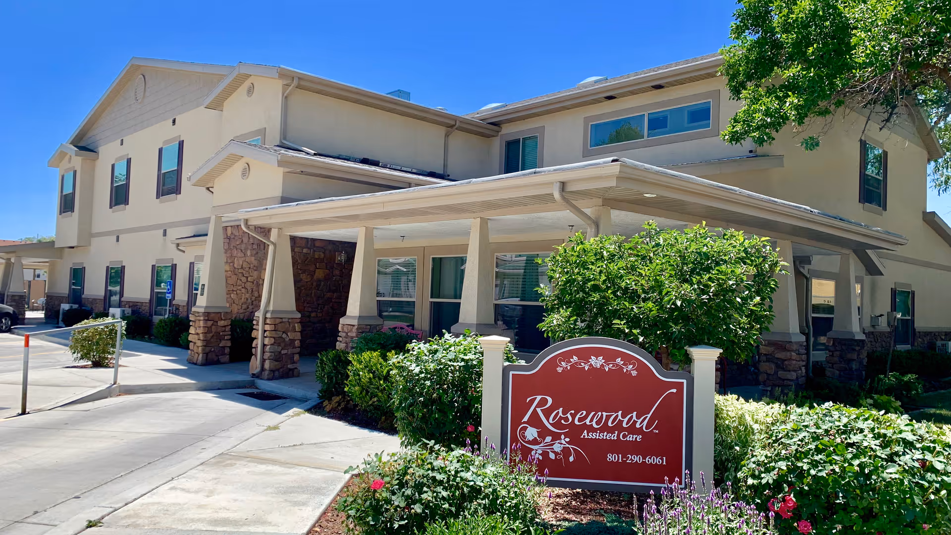 Exterior view of Rosewood Assisted Care facility showing a two-story beige building with stone accents and a covered entrance. There are bushes and flowers in front of the building, along with a red sign displaying the facility's name and phone number. The sky is clear and blue.