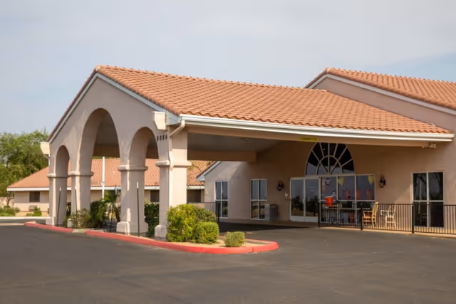 Exterior view of South Mountain Post Acute facility showing a beige stucco building with a red tile roof and an arched covered entrance. There are some bushes and plants near the entrance and a paved driveway in front.