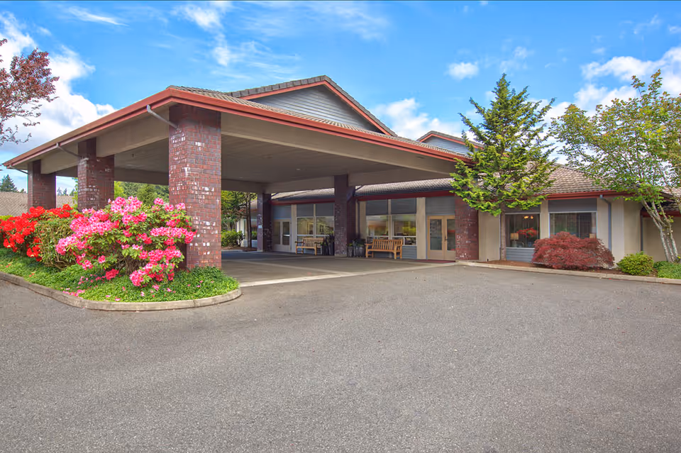 Covered entrance with brick pillars and flowering shrubs at the front of a single-story senior living building.