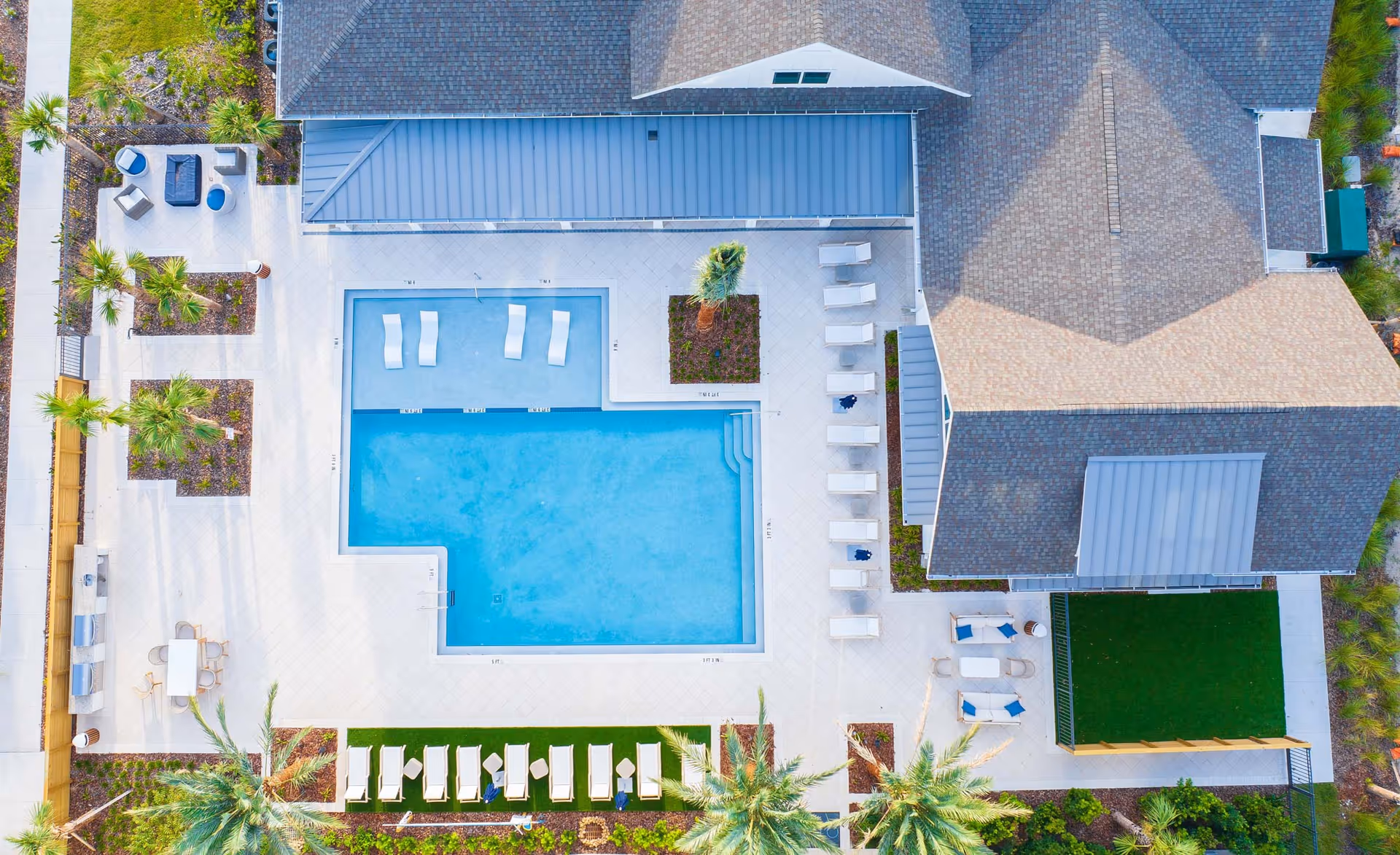Aerial view of a rectangular swimming pool with sun loungers, palm trees and surrounding buildings.