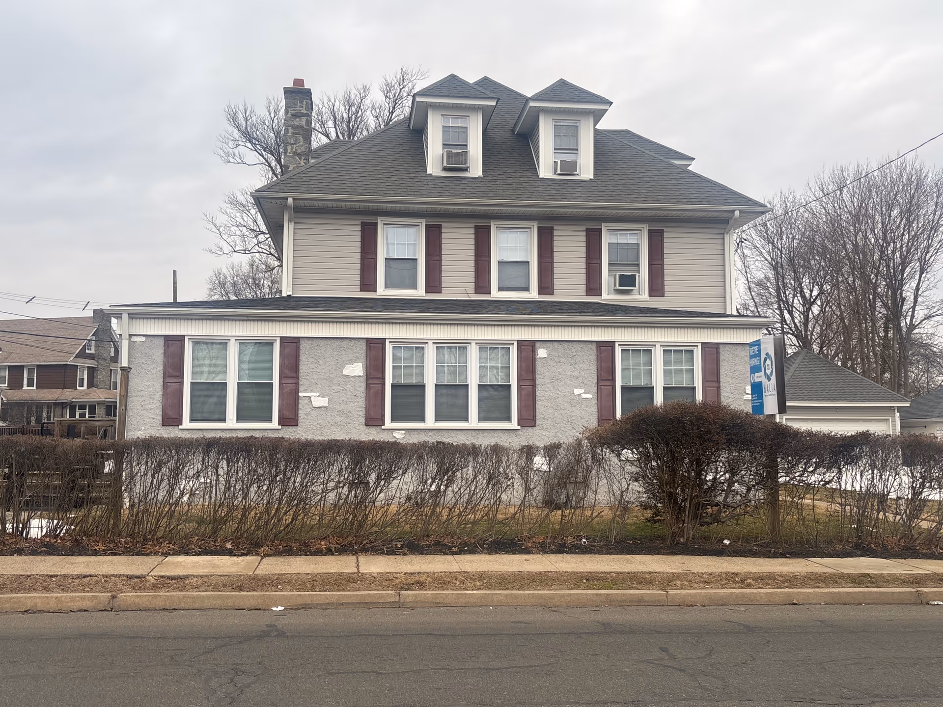 Gray two-story building with white trim, maroon shutters, and a sign near the sidewalk.