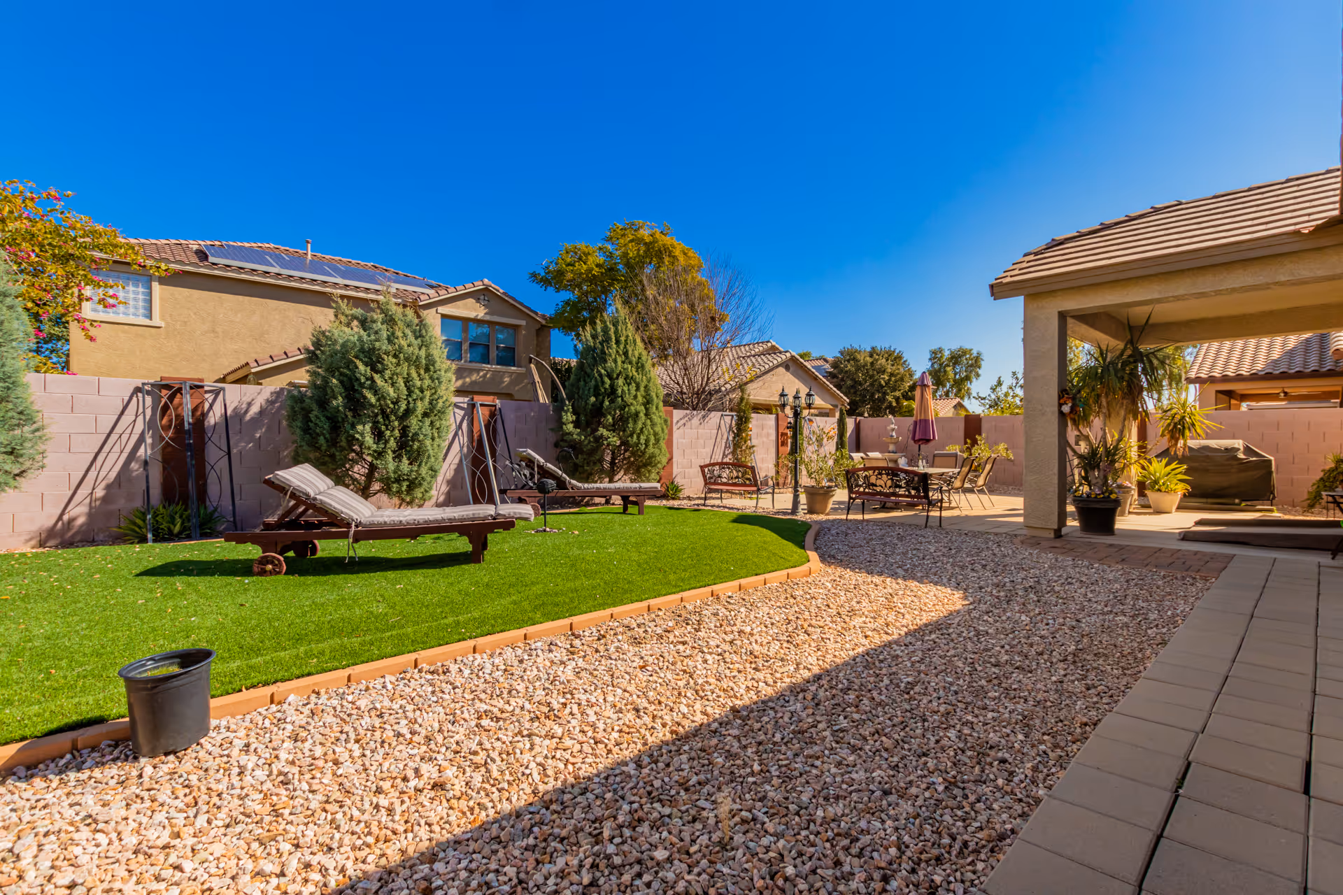 Sunny outdoor courtyard with lawn, lounge chairs, tables, potted plants, and a covered seating area.