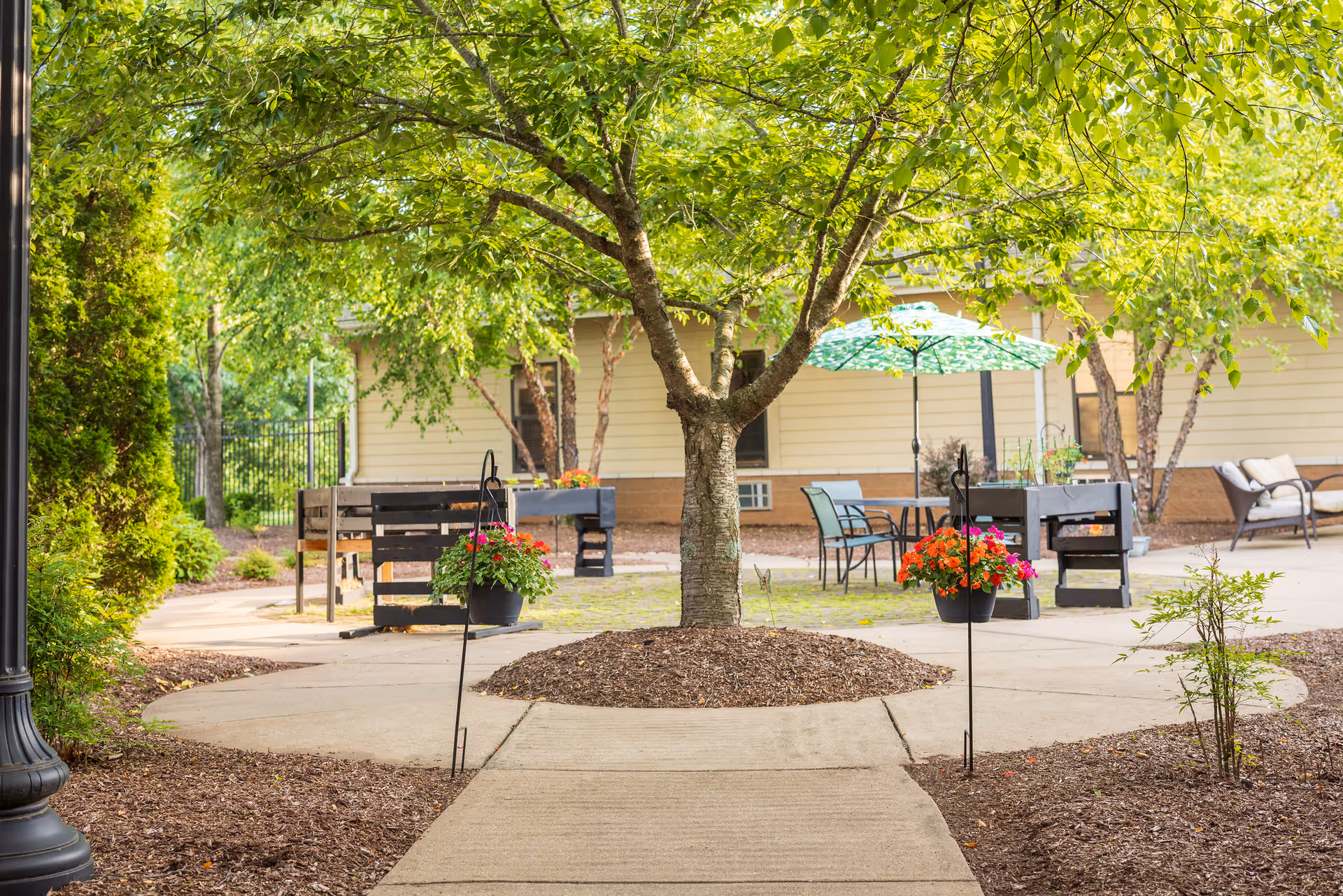 A peaceful outdoor patio area with a large tree in the center surrounded by a circular mulch bed. There are hanging flower pots with red and pink flowers on either side of the walkway leading to the tree. In the background, there are tables and chairs, some with a green patterned umbrella, and a beige building with windows. The area is shaded by trees and has a well-maintained garden with shrubs and mulch.
