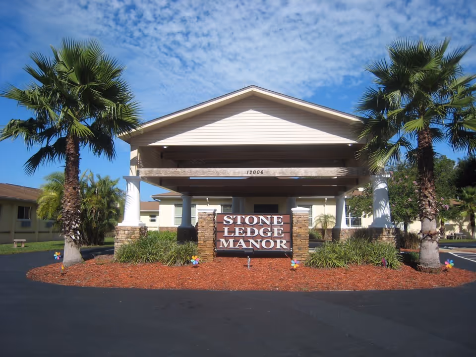 Entrance to Stone Ledge Manor Senior Living facility with a covered driveway supported by white columns and stone pillars, surrounded by palm trees and landscaping under a partly cloudy blue sky.