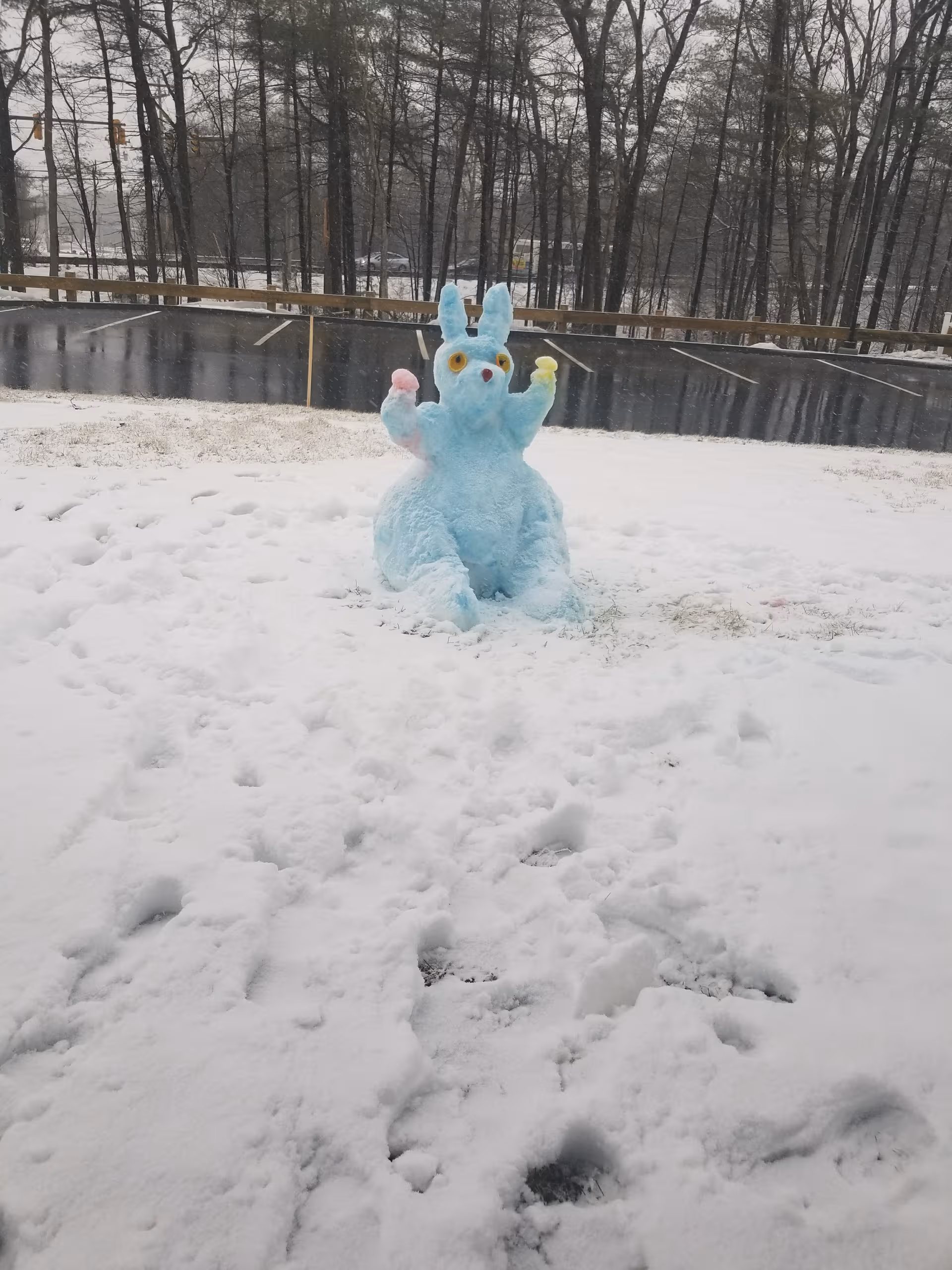 Blue snow rabbit sculpture sits on snow near a guardrail and bare trees.